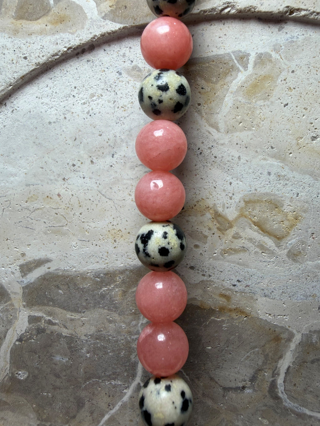 A close-up of a beaded strand featuring alternating pink polished round beads and black-and-white spotted round beads, laid vertically on a textured stone surface.