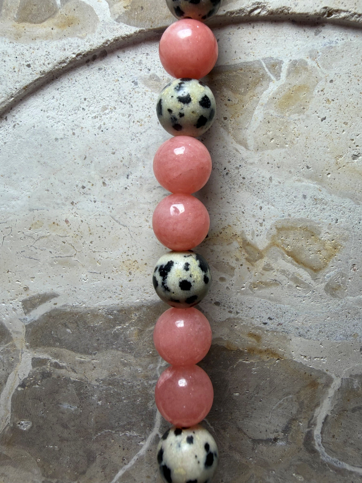 A close-up of a beaded strand featuring alternating pink polished round beads and black-and-white spotted round beads, laid vertically on a textured stone surface.